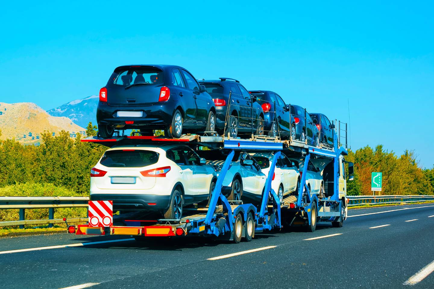 A car transporter truck carrying multiple automobiles, some black and some white, driving on a highway with trees and mountains in the background.