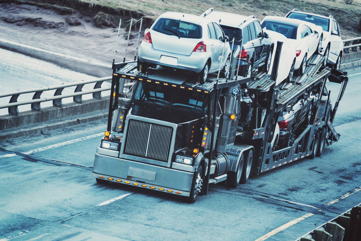 A transport truck carrying multiple automobiles is driving on a multi-lane highway near concrete barriers.