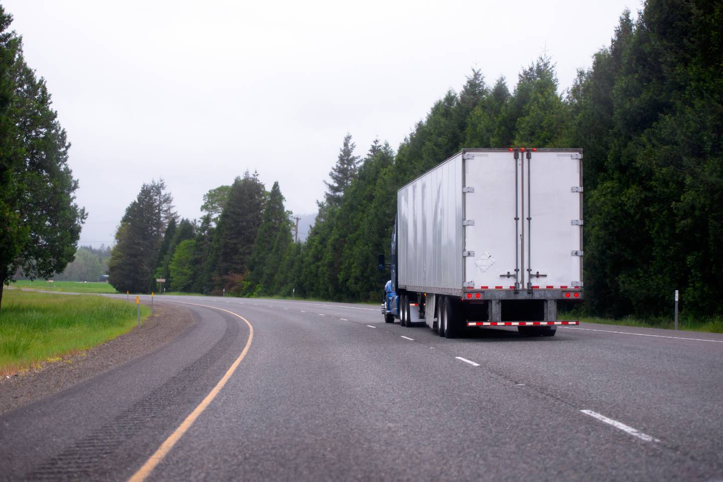 A white cargo truck drives down an empty, curved road surrounded by green trees, likely transporting goods in Dry Van Trailers.