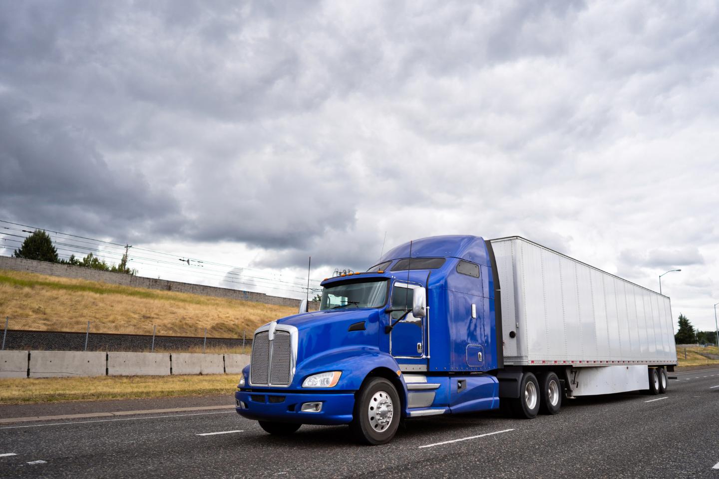 A blue semi-truck with a white dry van trailer drives on a paved road under a cloudy sky.