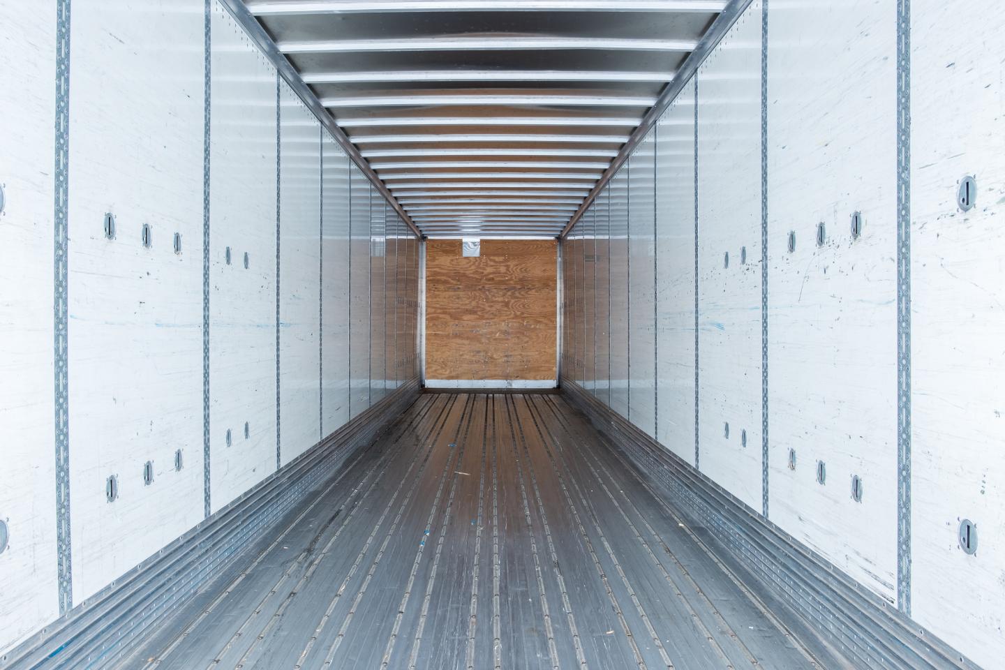 Interior view of an empty, white dry van trailer with metal floor and walls, and a wooden panel at the far end.