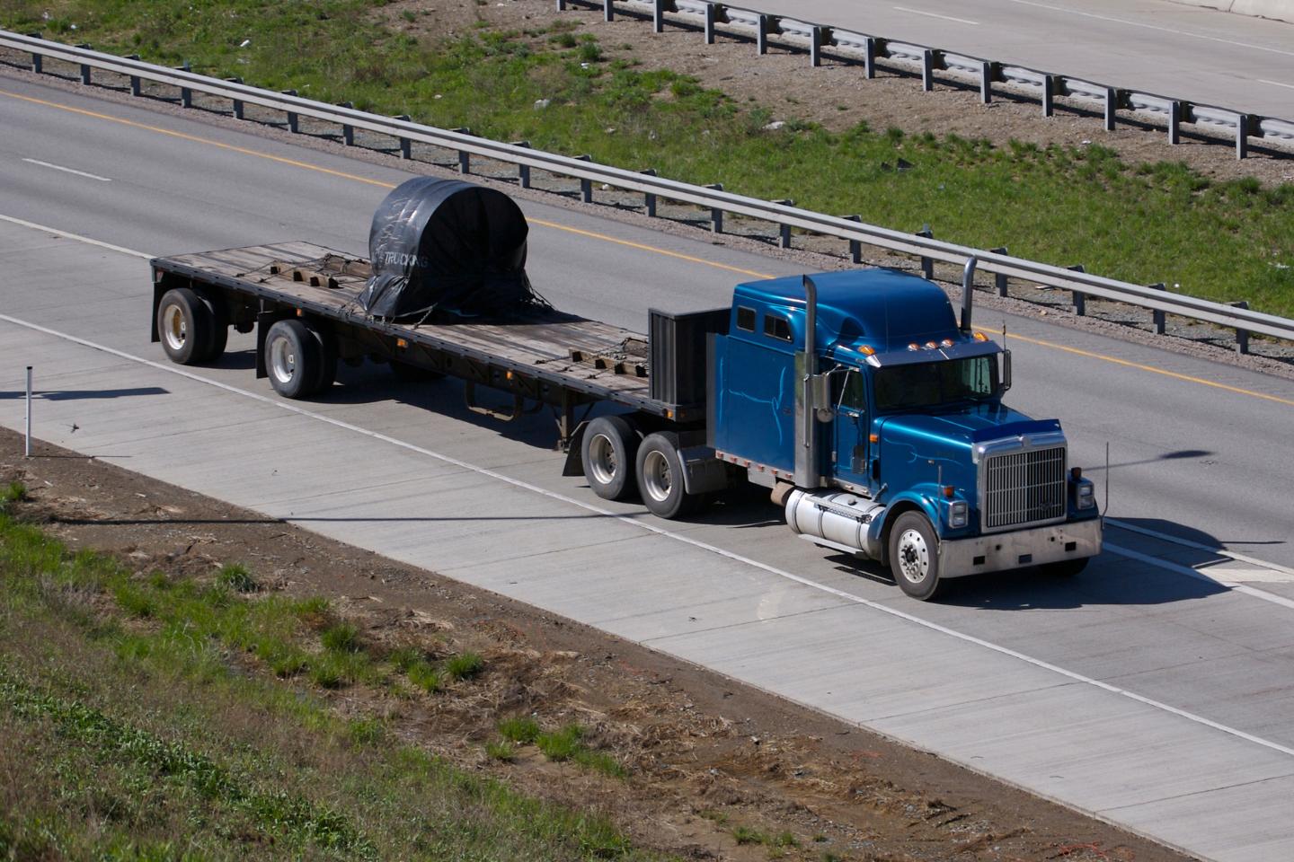 A blue semi-truck with a Step-Deck Transport trailer carries a large cylindrical load secured with a tarp, traveling on a multi-lane highway.