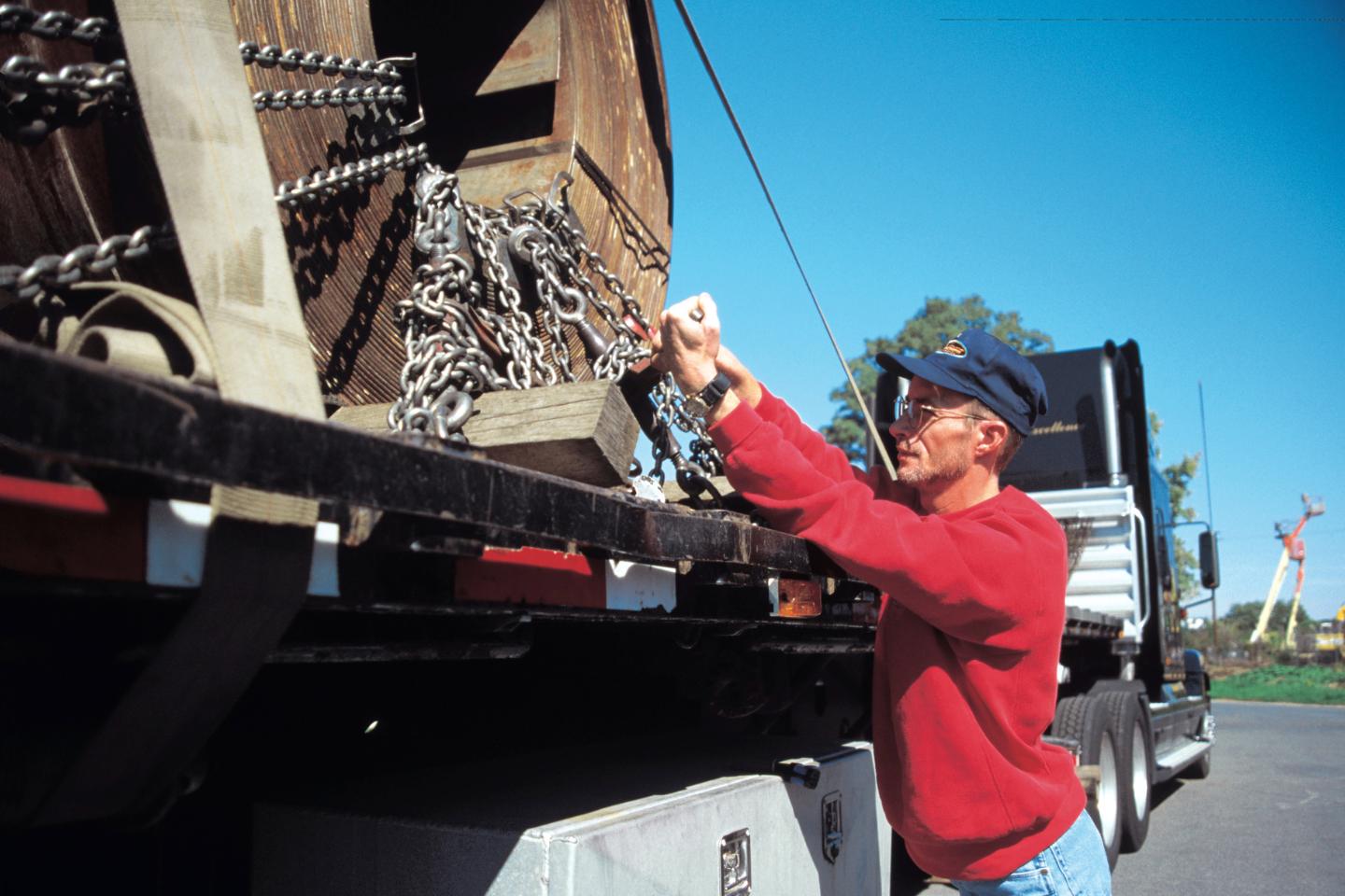 A man in a red sweater and cap secures chains around a large wooden spool on a step-deck transport truck.