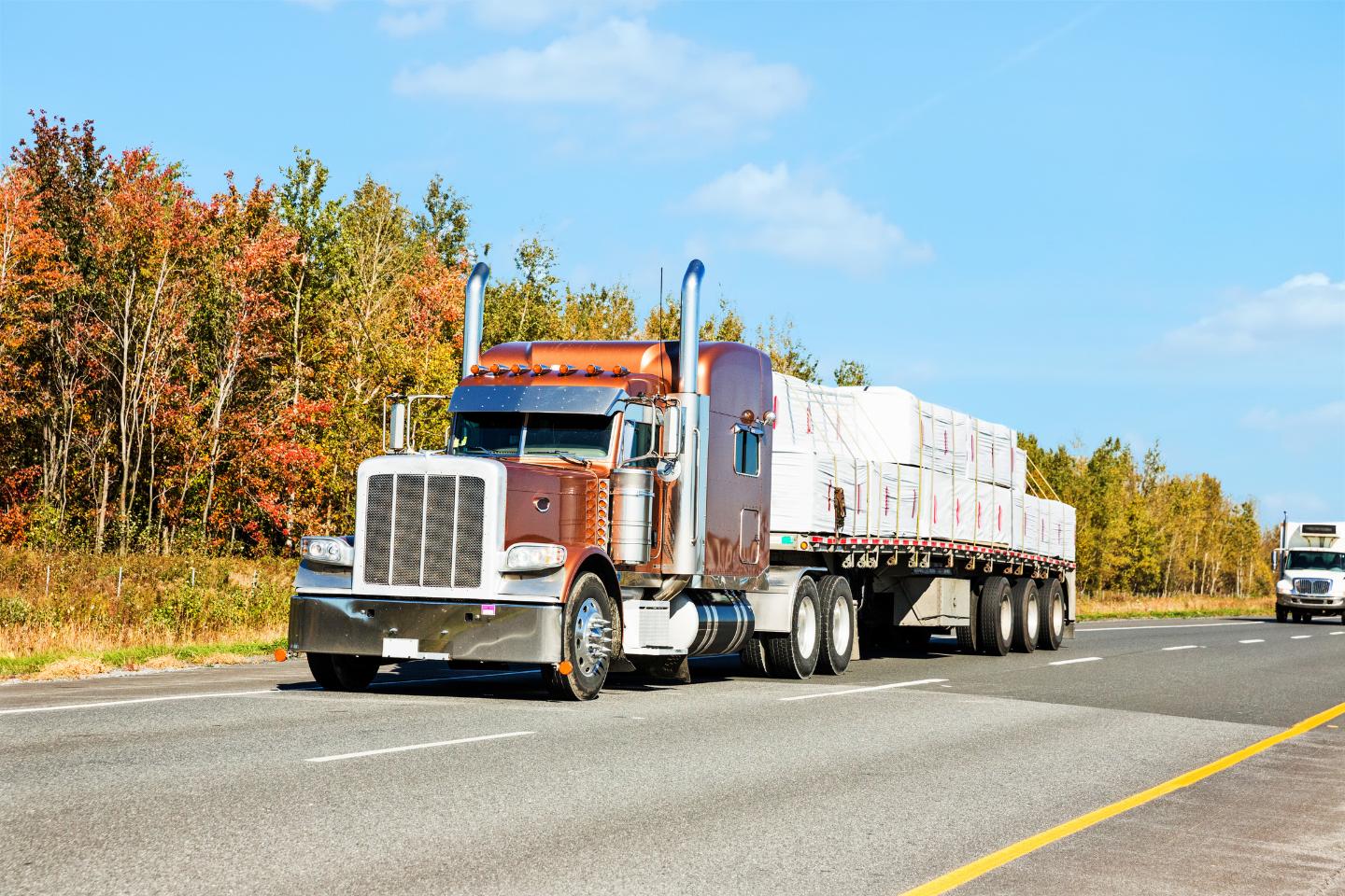 A large semi-truck with a step-deck transport trailer carrying stacked white packages drives on a highway beside a forest with autumn foliage.