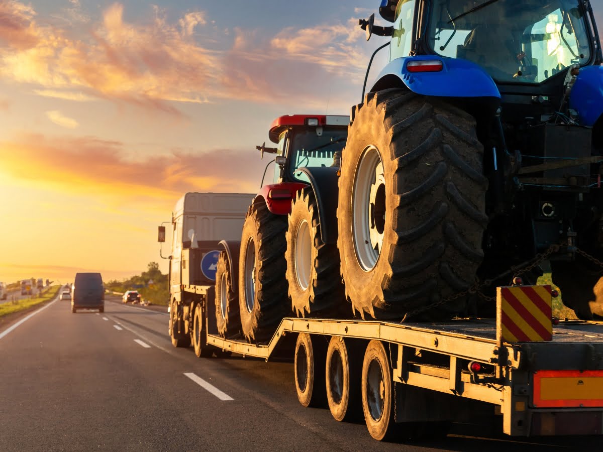 A truck providing hot shot delivery, transporting two large tractors on a flatbed trailer, drives along a highway at sunset, with other vehicles visible in the background.