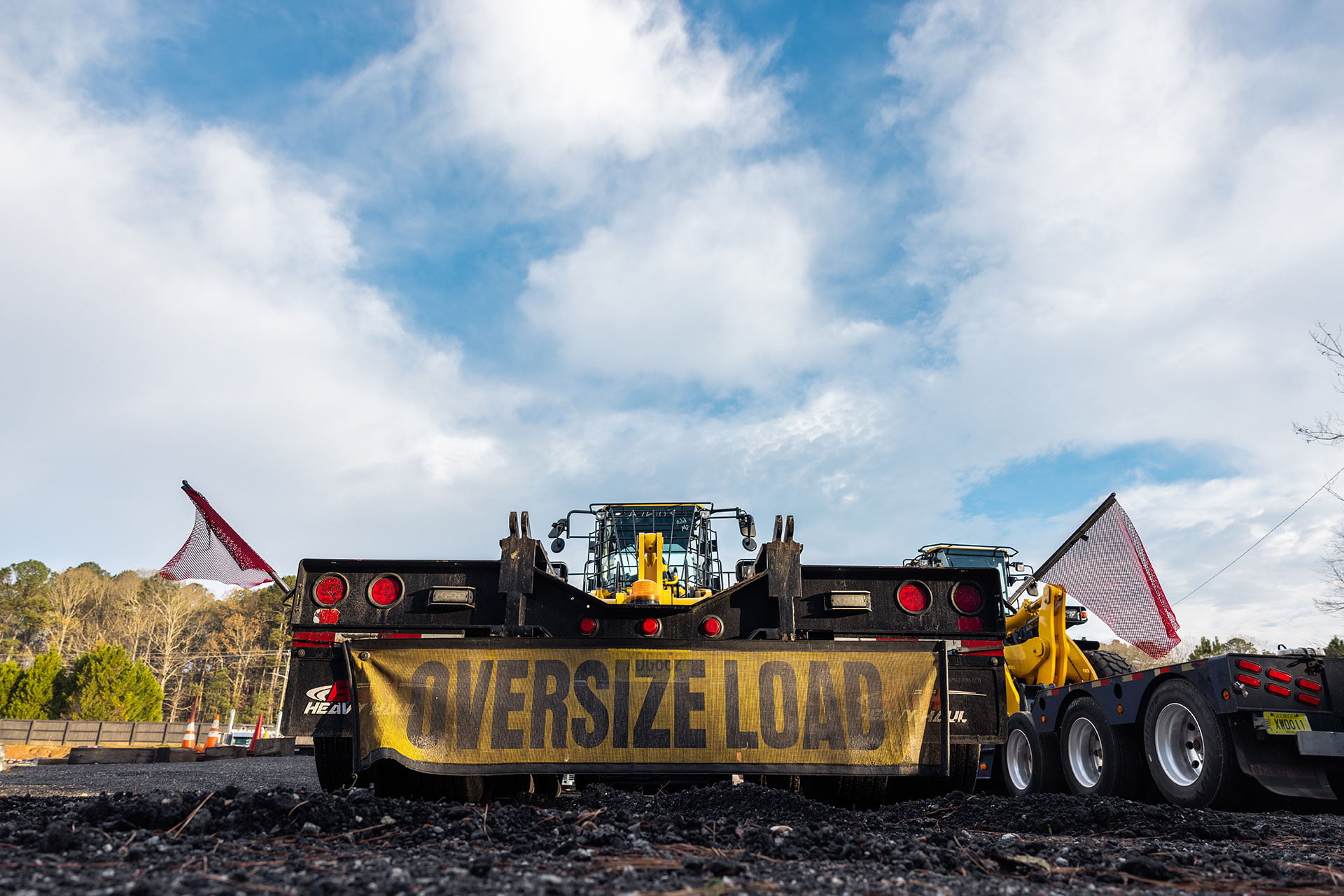 A large trailer labeled "OVERSIZE LOAD" is carrying industrial equipment, making its way to its new home. The sky is partly cloudy with some trees visible in the background.