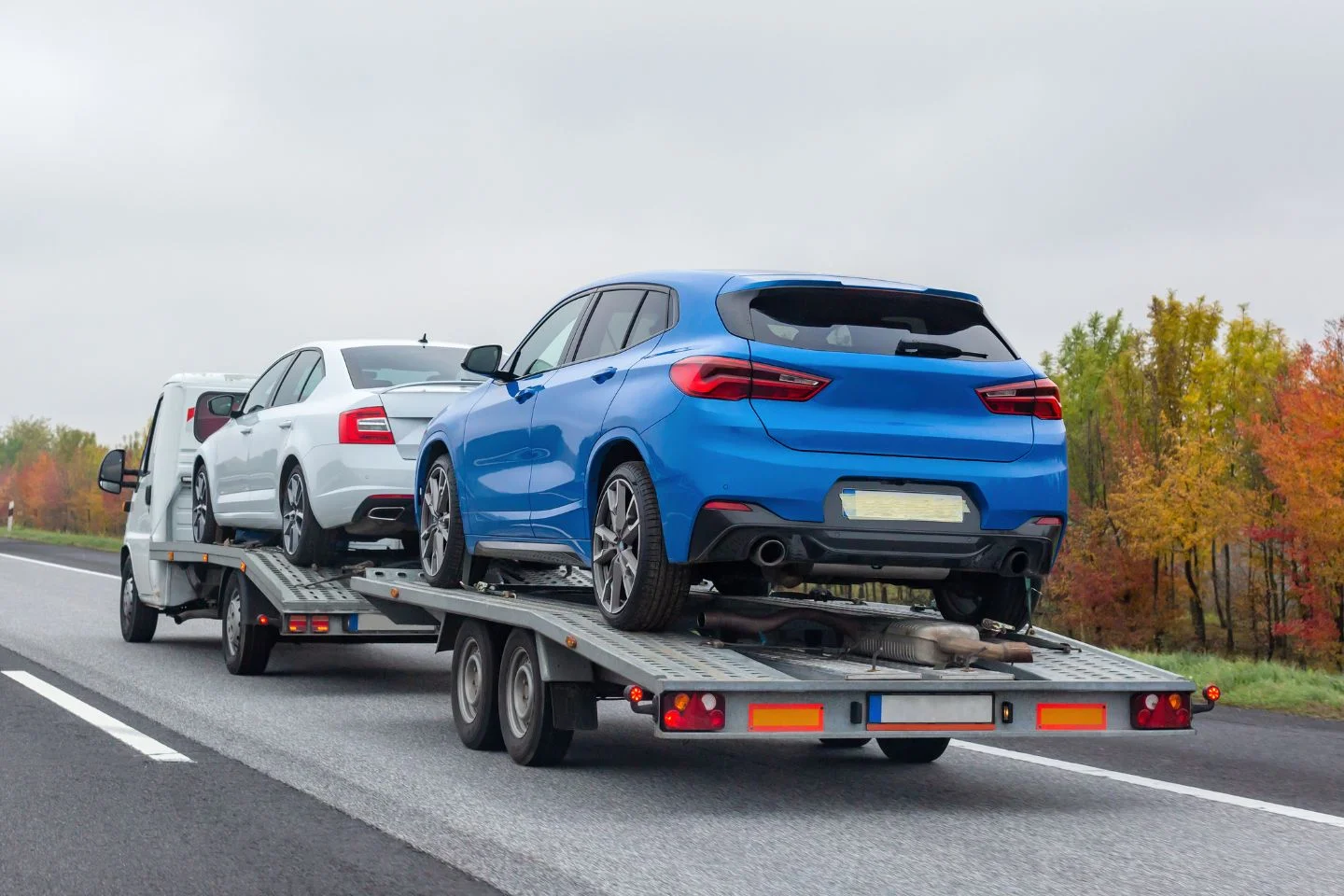 A blue car and a white car are being transported on the back of a flatbed tow truck, showcasing efficient logistics services, as it drives down the highway.