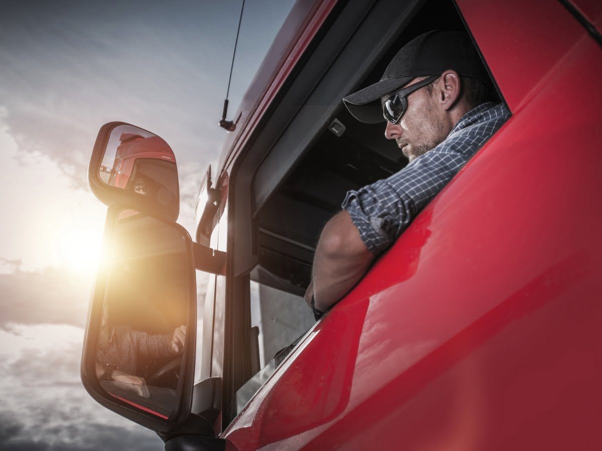 A truck driver wearing sunglasses and a cap looks out of the window of a red truck cab, captured on a sunny day, showcasing the essence of power-only trucking.