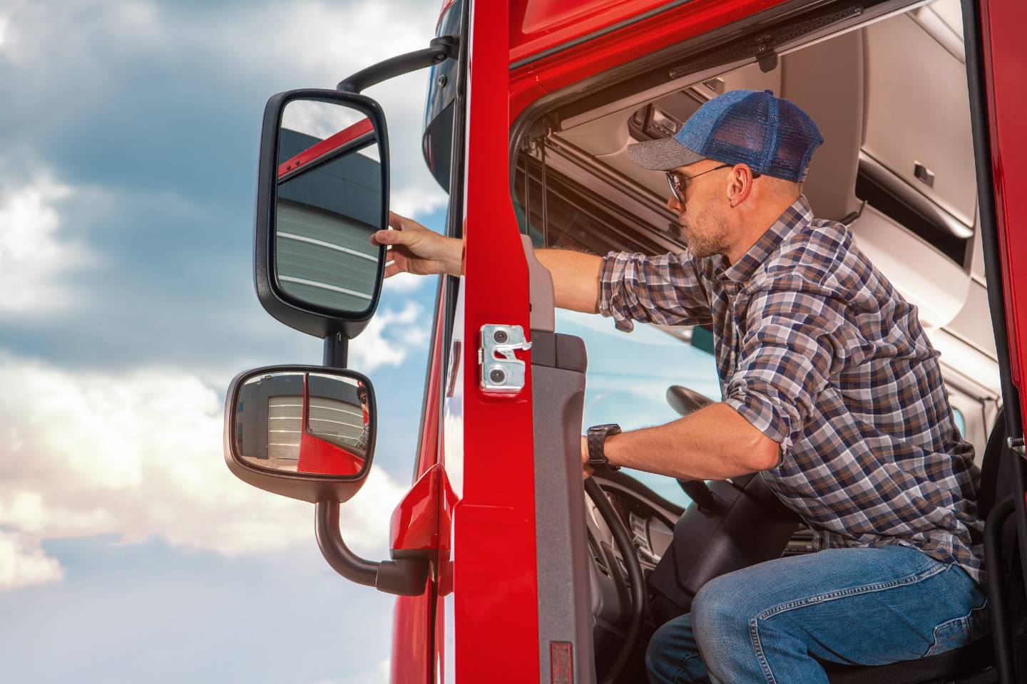 A person in a plaid shirt and cap adjusts the side mirror of a red truck, sitting in the driver's seat. The backdrop of a cloudy sky hints at the challenges and rewards of power-only trucking.