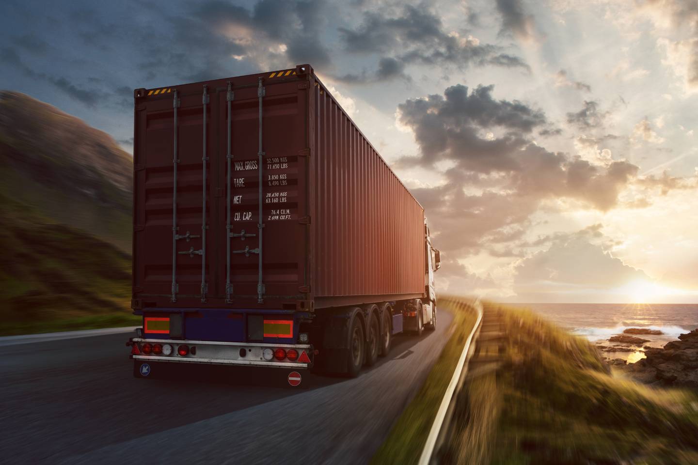 A large red power-only trucking container truck drives along a coastal road at sunset, with lush green hills on the left and the ocean on the right.