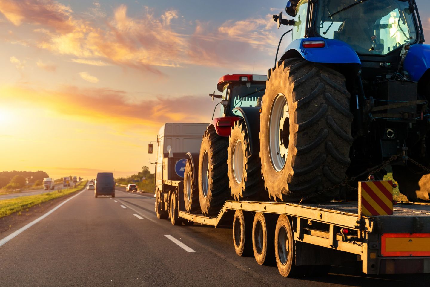 A large truck utilizes a step-deck transport to haul two blue tractors on a flatbed trailer down a highway at sunset.