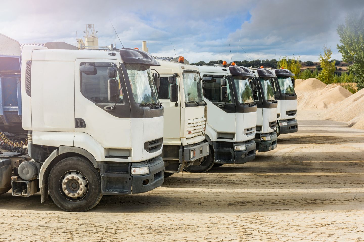 A row of five white trucks, specializing in Step-Deck Transport, parked on a sandy lot with mounds of sand and industrial buildings in the background.