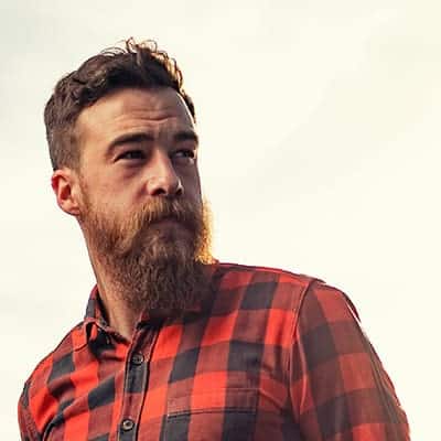 A man with a beard and mustache wearing a red and black plaid shirt looks away from the camera, against a light background.