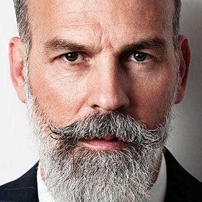 Close-up portrait of a serious-looking man with a full grey beard and mustache, wearing a dark jacket and white shirt against a light background.