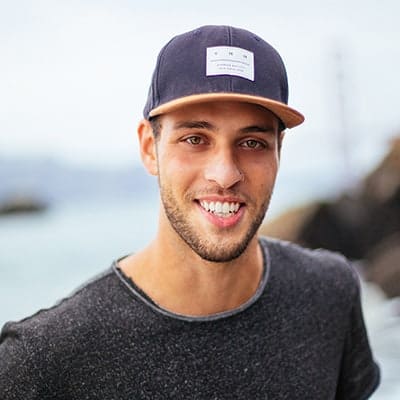 A man wearing a dark-colored baseball cap and a gray shirt smiles at the camera. The background is an outdoor scene, possibly near a body of water.