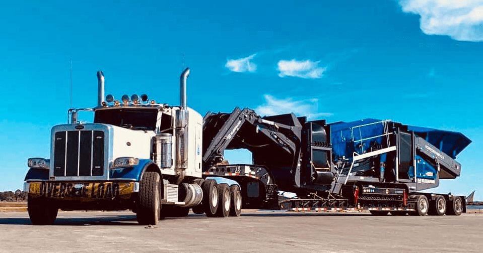 A large white semi-truck from KLR Trucking is towing a massive piece of blue industrial machinery along a wide, flat roadway under a clear blue sky.