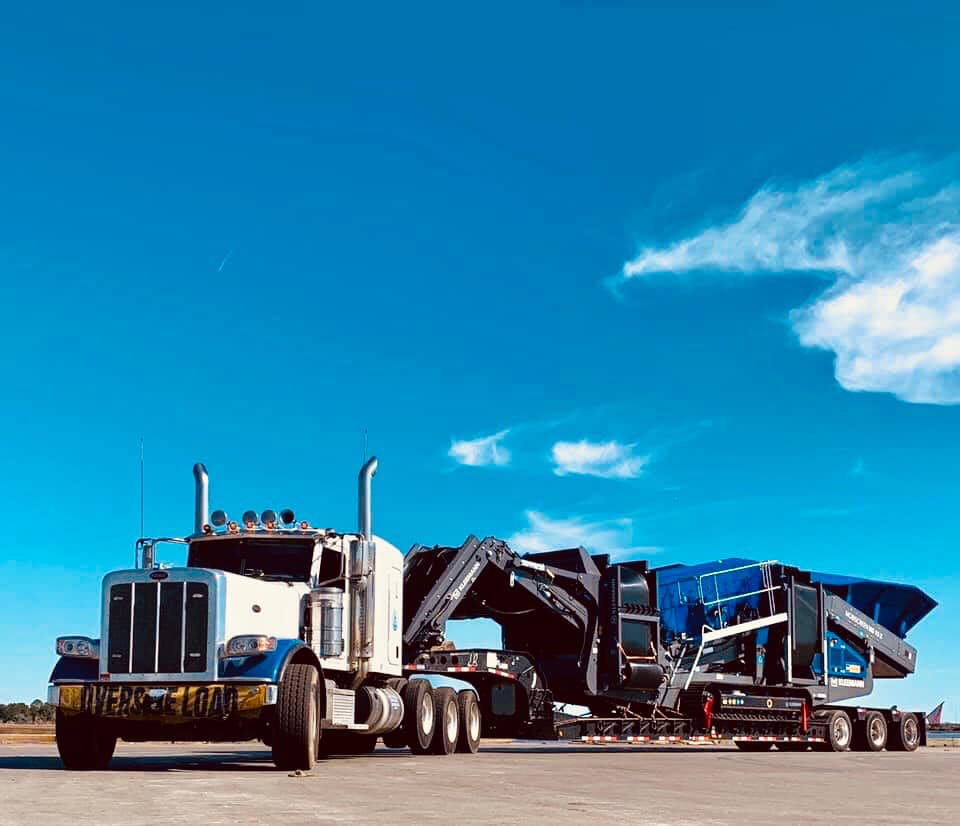 A large white semi-truck for about KLR Trucking with a flatbed trailer transports heavy blue machinery on a clear, sunny day, moving efficiently towards its destination to support the construction of new homes.